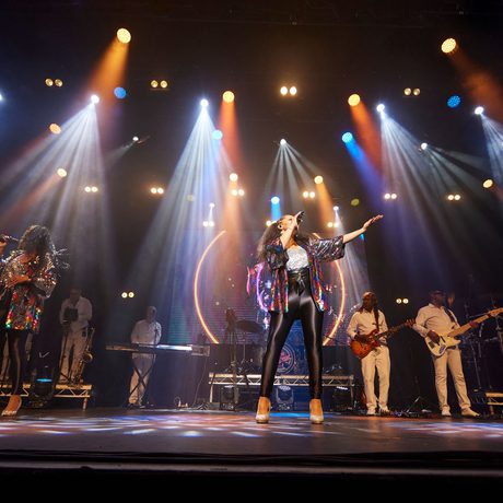 Three performers dressed in rainbow sequined jackets sing while the band, dressed in white, stand behind
