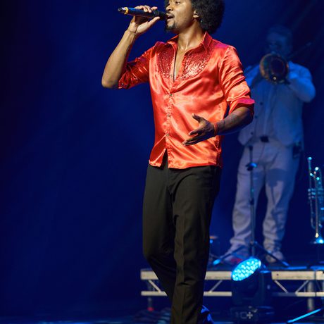 man in red silk shirt and black trousers sings into microphone