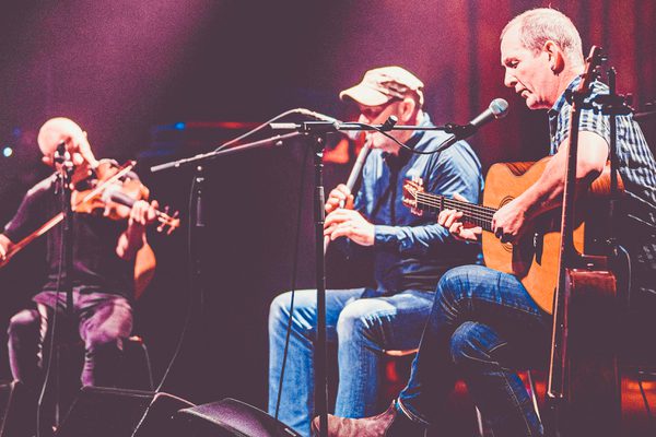A violinist, guitarist and whistle player sit in front of microphones