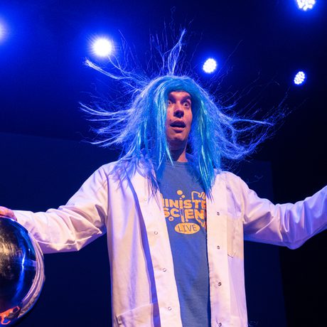 A person in a lab coat and a blue wig, with one hand on a big metal ball.