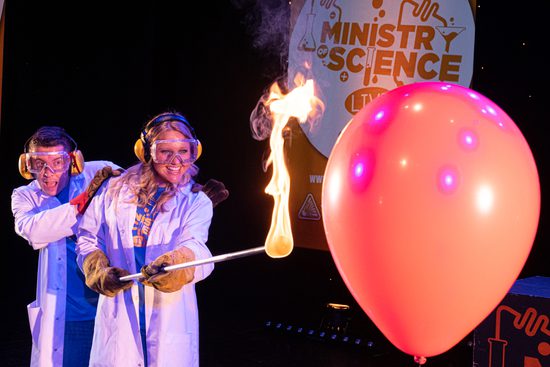 Two people in lab coats, ear defenders and goggle holding a flame towards a big red balloon.