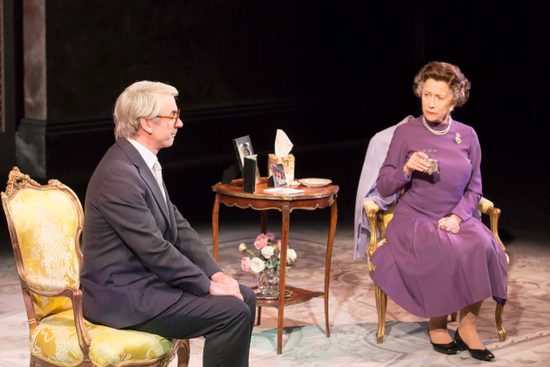 A woman and a purple dress and a man in a grey suit have a conversation. They are both sat on yellow chairs.