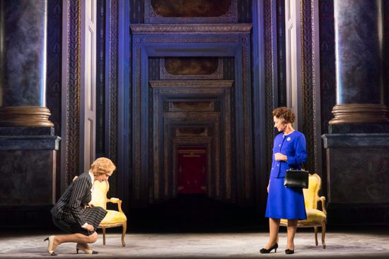 A woman in a black and white pinstripe suit curtseys towards a woman in a vibrant blue suit.