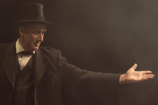 A man in a black coat and black top hat reaching one hand out to the side.