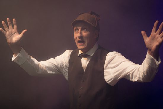 A man wearing a deerstalker hat and a waistcoat with his hands in the air, looking scared.
