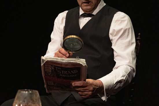 A man standing at a desk wearing a waistcoat, looking at a newspaper through a magnifying glass.