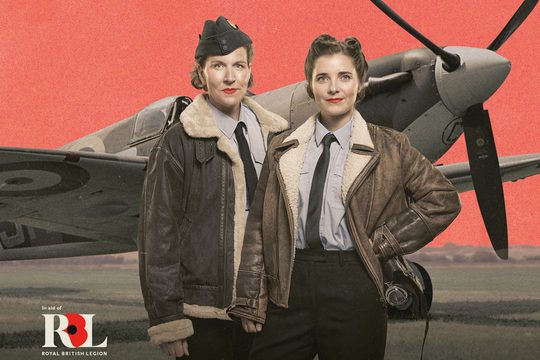 Two women dressed in fleece lined leather jackets stand in front of a spitfire