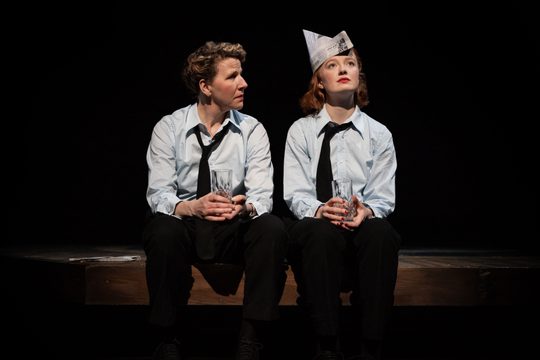 Two women in pale shirts and black ties sit on a dark stage holding glass tumblers. One wears a newspaper hat looking into the distance while the other looks at her.