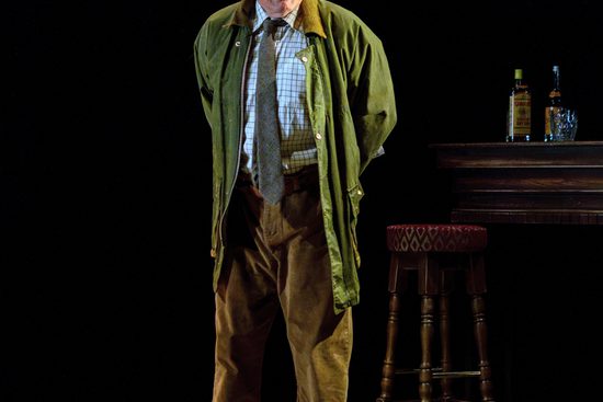 Man in green jacket and blue checked shirt stands with his hands behind his back