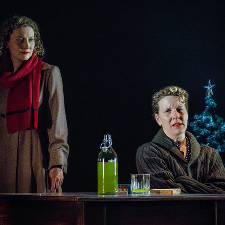 Woman in red scarf looks towards another woman sat at a table with a bottle of florescent liquid in front of her
