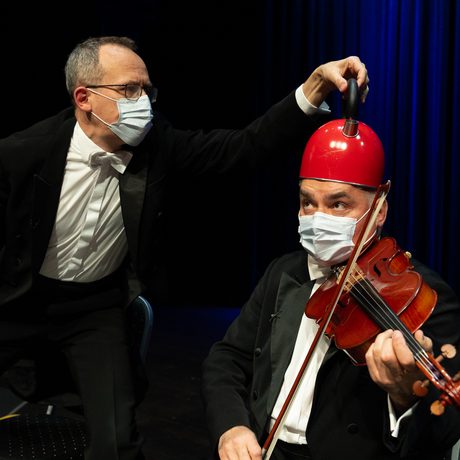 A man playing the viola while another man holds a red kettle on his head.