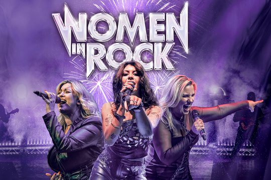 Three women singing into micro phones under purple lights, behind them fireworks go off.