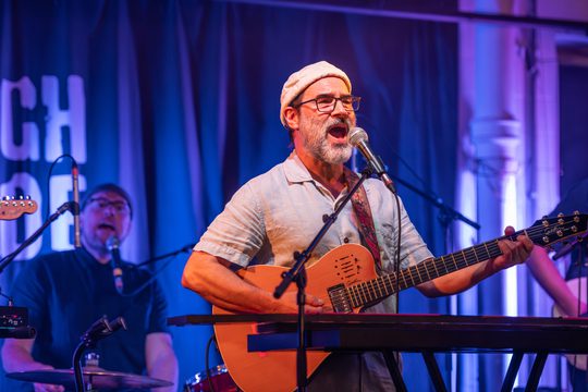 A man in a shirt and a hat plays an orange guitar on a stage. He is also singing into a microphone.