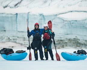 A man and a woman stand holding paddles next to kayaks in the arctic.