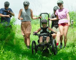 Five people wearing helmets. Four of them are standing and helping the other person, who is in an adapted bicycle, to cycle through long grass.