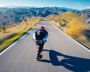Someone on a skateboard going fast downhill. They are bent forwards, and the scenery around them is beautiful mountains.