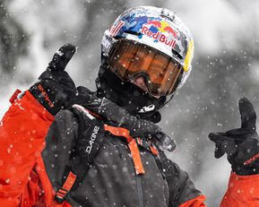 A person in a red ski suit and helmet with the red bull logo on it pointing to the sky.