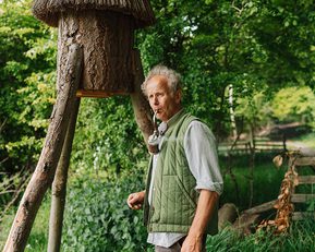 An older man in a green jacket stands in a woodland next to a wooden beehive.