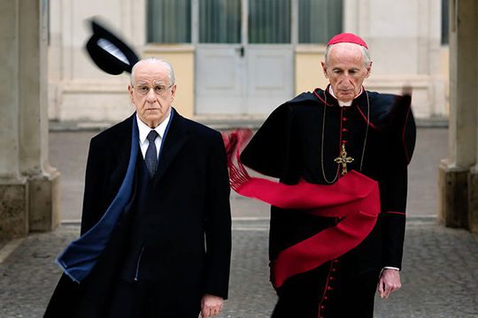 A man in a black suit with a blue scarf's hat has been blown off by the wind. He is walking with another man in black garments, a red scarf, a red hat, and a cross around his neck.