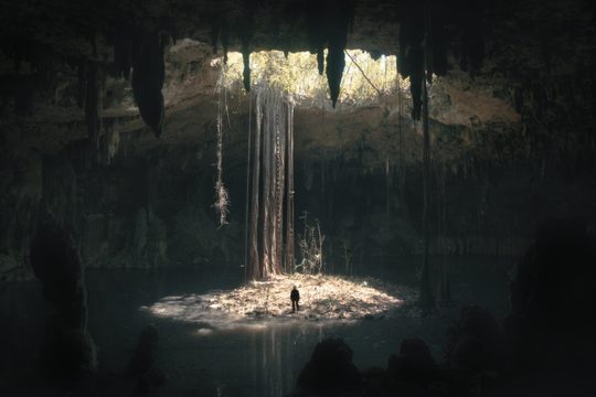 A large cave lit through a hole in the roof. A person looks tiny inside it to show scale.