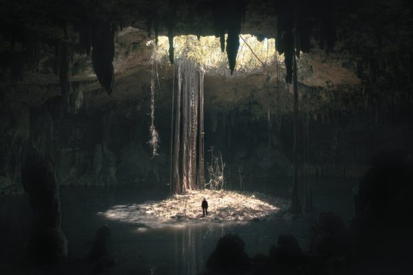 A large cave lit through a hole in the roof. A person looks tiny inside it to show scale.