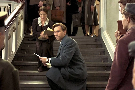 A man in a long grey coat crouches on a staircase holding some papers. People at the top and bottom of the stairs are all looking at him.