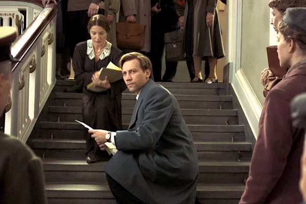 A man in a long grey coat crouches on a staircase holding some papers. People at the top and bottom of the stairs are all looking at him.