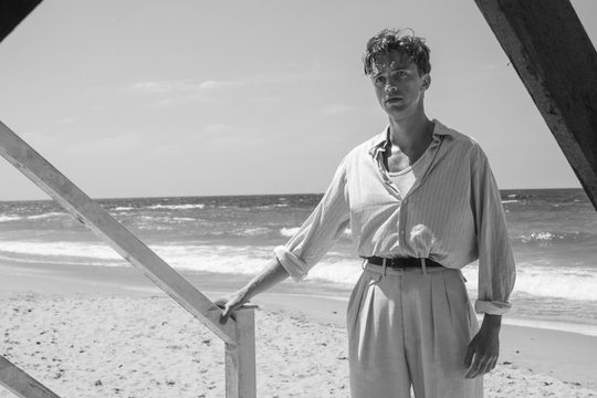 Black and white image of a man in a shirt with the sleeves rolled up, tailored trousers and a belt. He is standing on a beach.