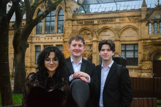 Two men and one woman wearing black. They are carrying instruments in cases, and standing outside an impressive stone building.