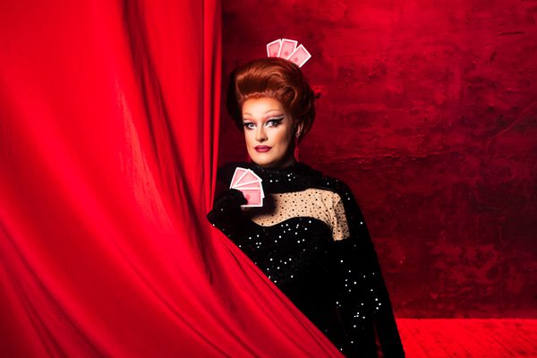 Kate Butch in a black sparkly dress, draped in a red curtain and holding red playing cards.