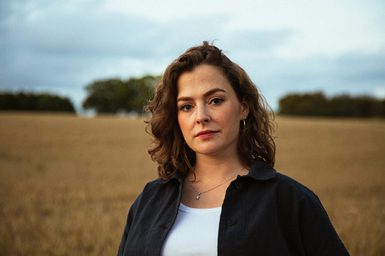 A lady with brunette curly hair, a white T-shirt and a black overshirt standing in a wheat field.