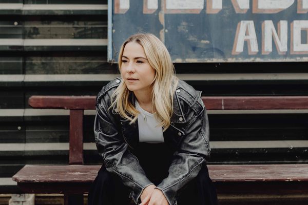 A woman with blonde hair wears a leather jacket and sits on a brown bench.