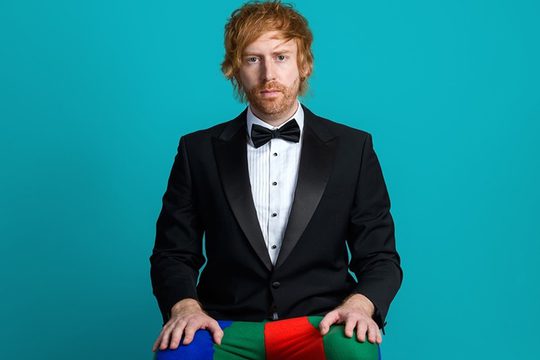 Man in suit jackets and clown trousers sits in front of turquoise background