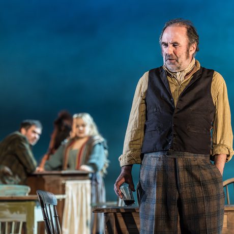 A man in a waistcoat and tweed jacket holds the end of a pint of beer in his hand. He is in a pub.