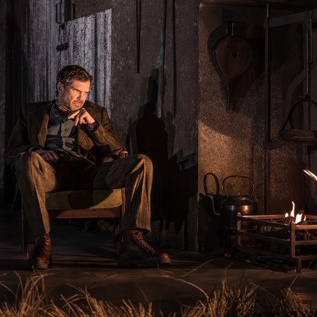 A grumpy man sits in a wooden chair by a dwindling fire.