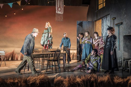 A group of people in colourful clothing gather around a man sat on the floor. They all look angry.