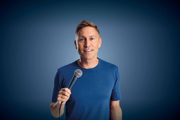 A blonde man stands in a blue t-shirt holding a microphone.