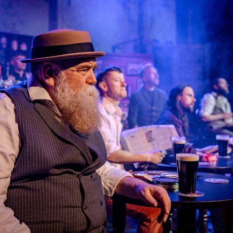 An elderly man in a straw hat and a waistcoat holding a walking stick sits in a pub.