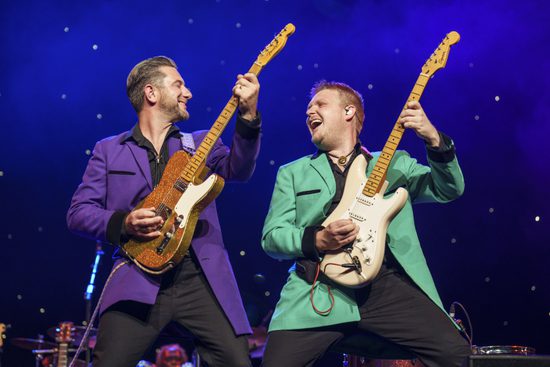 Two guitarist, one in a purple jacket and one in a green jacket hold up their guitars and smile at each other