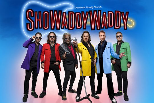 6 members in different coloured jackets stand in front of one microphone