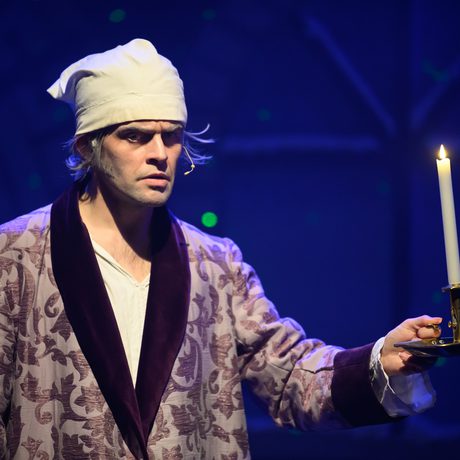 A man in a purple nightgown and white hat holds a candle out in front of him.