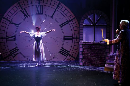 A woman in a white flowing dress stands in front of a large clock with cracks in it. A man is holding a candle to her and looking afraid.