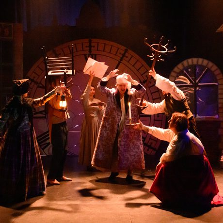 A man wearing a purple nightgown looks frustrated, and is surrounded by people holding various objects, including a chair, a book, and a coatstand.