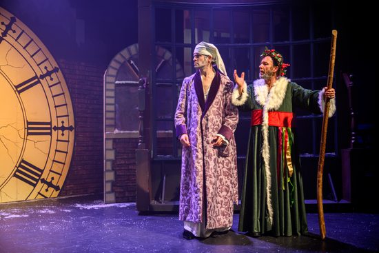 A man wearing a purple nightgown and a white hat is talking to another man wearing a green cloak and a crown made of holly.