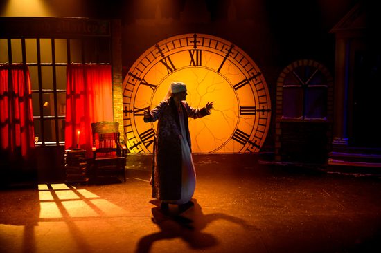 A man in a long nightgown stands in the glow of a large clock, lit in warm orange.