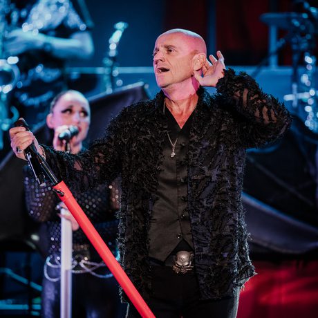 A man in black clothing. He had one hand holding a red microphone stand, and one hand raised to his ear.