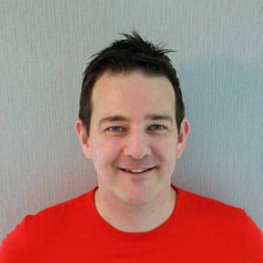 A white brunette man in a bright red t-shirt smiles.