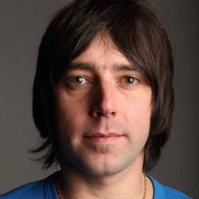 A white man with jaw-length brown hair in a blue t-shirt looks straight into the camera.
