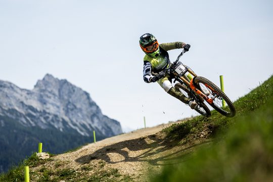 A man in a green biking suit riding through grass on beautiful mountains.