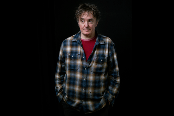 Dylan Moran a white man with messy hair stands with his hands in his pockets wearing a checked shirt and a red t-shirt underneath.